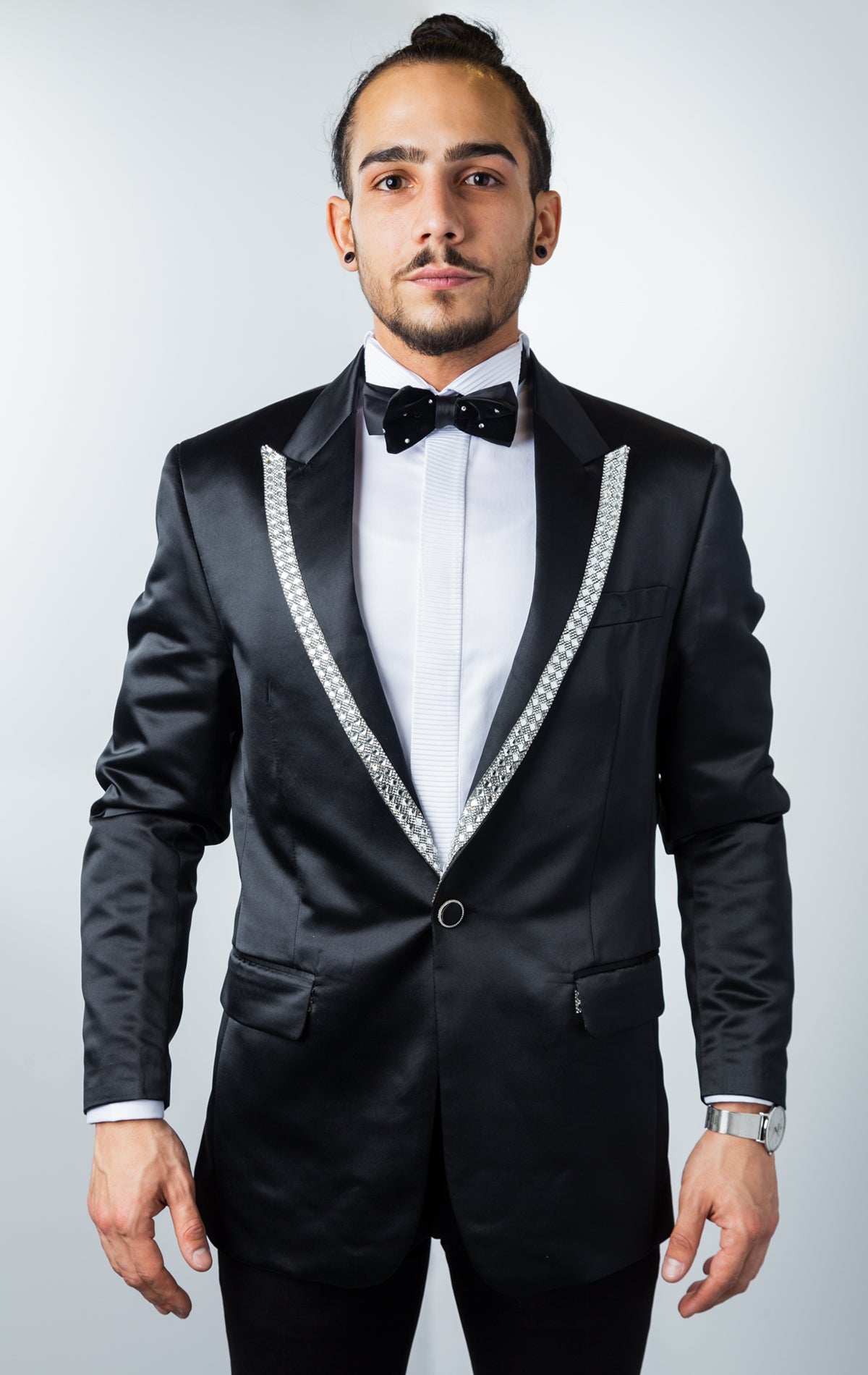 Black, one-button tuxedo jacket with a slim fit. The jacket features a shiny black fabric, rhinestone embellishments on the collar, a pocket square, and button cuffs. The jacket is made from a medium-lightweight fabric.